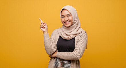 Smiling young Asian Muslim woman in hijab pointing up at copy space, isolated on a yellow background.