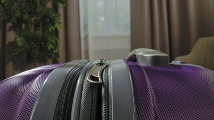 Detailed view of a purple suitcase with a metallic handle and zipper, in a room with plants and curtains.