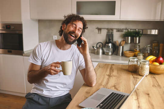 A man sits in a modern kitchen, smiling as he talks on the phone, holding a coffee mug in one hand and a laptop open on the kitchen counter beside him.