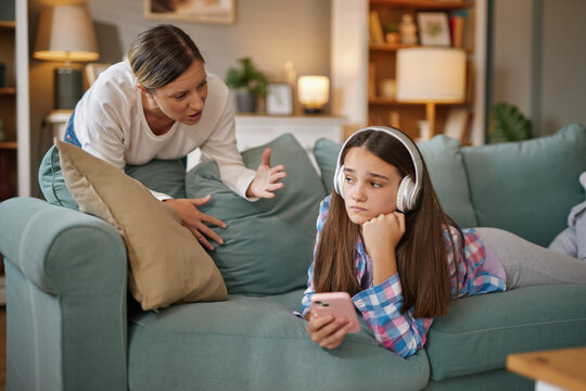 A teenage girl relaxes on a sofa, wearing headphones and focused on her phone, as her mother talks animatedly, conveying worry in a warm living room setting.
