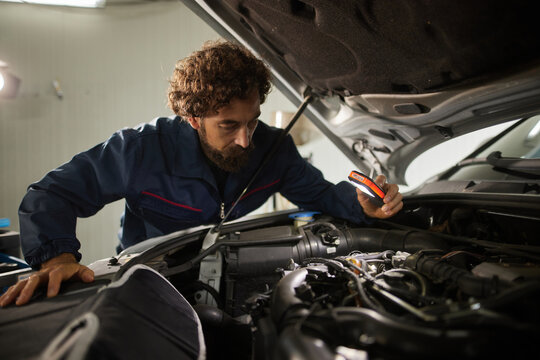 A skilled car mechanic works under the hood of a vehicle, carefully checking the engine parts for any issues. The workshop is well-lit and organized.