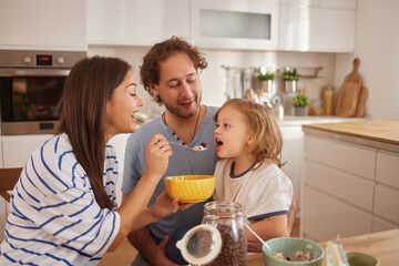 In a bright kitchen, a woman feeds a young child from a yellow bowl while a man smiles at them, highlighting a joyful family moment during breakfast.