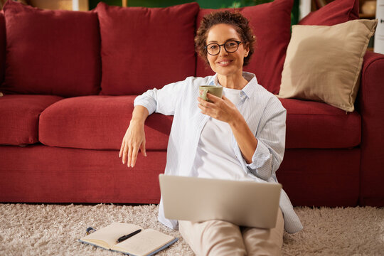 A woman sits on a soft rug in her living room, sipping coffee and working on her laptop. She appears relaxed, with books scattered around, enjoying her work-from-home setup. - Powered by Adobe