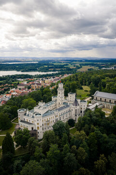 Aerial view of the striking white Zamek Hlubok&aacute; castle amidst a sea of verdant trees under a vast, cloudy sky, Hluboka nad Vltavou, Jihocesky kraj, Czechia.
