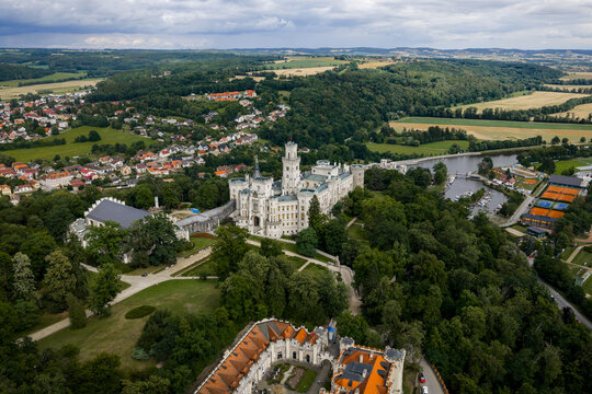 Aerial view of the gleaming white Zamek castle amidst lush green trees, contrasting with the town and fields beyond, Hluboka nad Vltavou, Jihocesky kraj, Czechia.