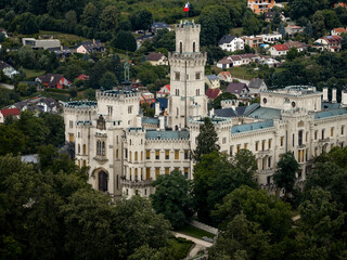 Aerial view of the creamy white Zamek castle, crowned with the Czech flag, rises majestically amidst a sea of green trees, Hluboka nad Vltavou, Jihocesky kraj, Czechia.