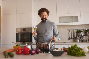 A smiling man stirs a pot on the stove in a sleek kitchen, holding a glass of white wine. Fresh vegetables and herbs are spread across the counter, creating an inviting cooking atmosphere.