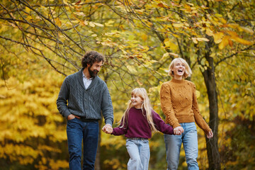 A family takes a joyful stroll through a park filled with autumn leaves. They share laughter and hold hands, enjoying their weekend together in the crisp air under colorful trees.