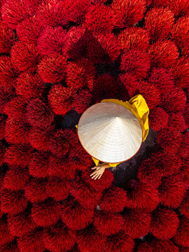 Aerial view of a person in traditional conical hat surrounded by vibrant red incense sticks, creating a striking contrast of color and texture, Quang Phu Cau, Hanoi, Vietnam.