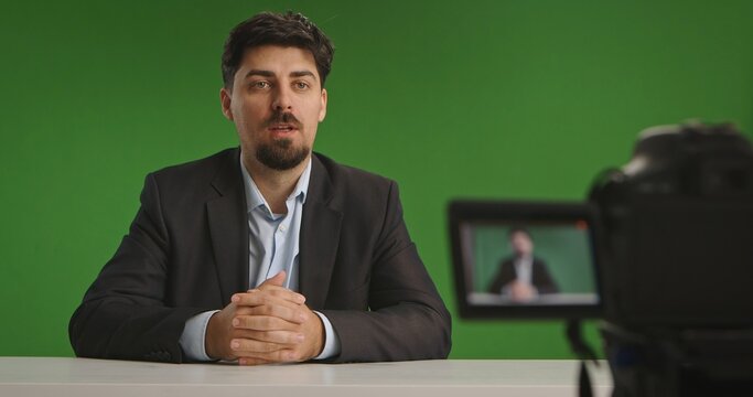 Man in a suit sitting at a desk in a live streaming studio with a camera and green screen backdrop.