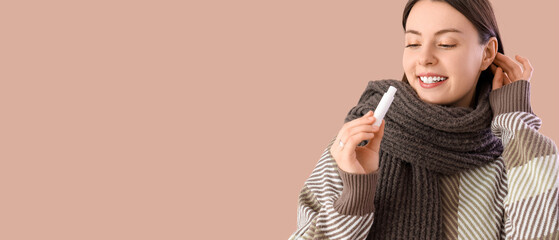 Young woman in winter clothes with lip balm on beige background, closeup