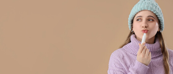 Woman in winter clothes applying lip balm on brown background
