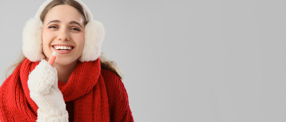 Happy woman in winter clothes and with lip balm on grey background