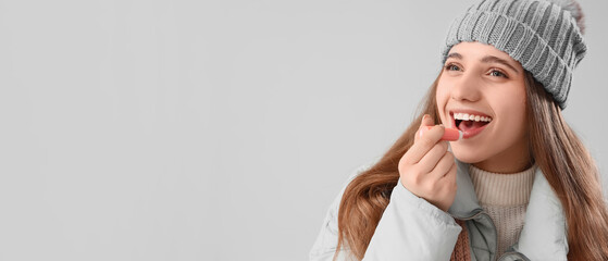 Happy woman in winter clothes applying lip balm on grey background