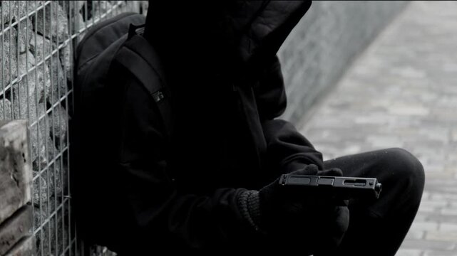unrecognizable killer, bandit, or man in a hood and mask leaning against a wall, in ambush, reloading a pistol magazine and firing a shot, armed assault, robbery