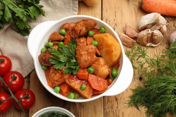 Delicious cooked stew in bowl and ingredients on wooden table, flat lay