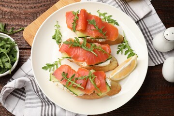 Tasty bruschettas with salmon, cream cheese, cucumber, arugula and lemon on wooden table, flat lay