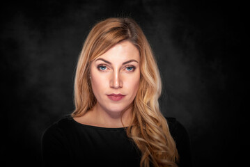 Fototapeta premium Portrait of a confident blonde woman with wavy hair wearing a black top against a dark studio background, looking directly at the camera with a serious expression.