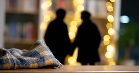 Silhouetted couple holding hands with warm bokeh lights in cozy indoor setting and folded checkered blanket in foreground