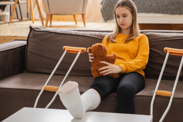 Young girl with broken leg cast sitting on sofa. Sad little kid hugging her teddy bear recovering from broken leg at home. Girl using cast and crutches. Accident injury fracture treatment foot bandage