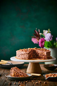 a side angle of a decadent chocolate mocha cake sprinkled with hazelnuts on a marble cake stand with a slice served in the foreground, against a rich, moody custom green backdrop.