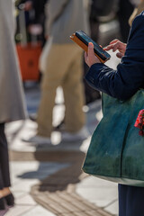 Business person in blue jacket using smartphone with brown leather case while holding green patina tote on crowded city street corner during morning rush.
