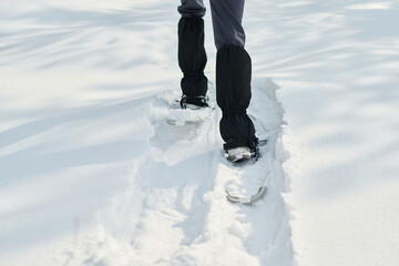 Young adult Caucasian woman walking through deep snow wearing snowshoes, lower legs and feet visible, leaving clear tracks in fresh snow, outdoor winter activity in progress
