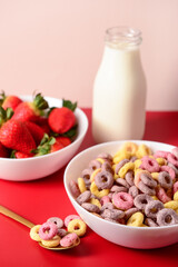 Bowls with colorful sweet cereal rings, strawberries and bottle of milk on red table