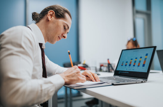 A focused businessman wearing a white shirt and tie writes in a notebook while reviewing colorful bar charts on a laptop screen in a bright corporate office.