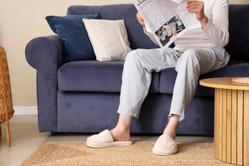Man in soft slippers resting on sofa and reading newspaper at home