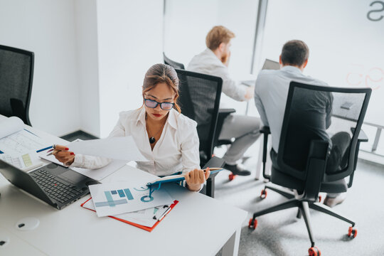 A focused businesswoman at a modern office reviews papers beside a laptop, while colleagues work in the background.