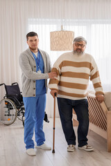 Young caregiver taking care of senior man with stick at home