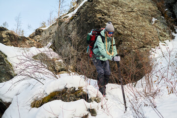 Caucasian young adult man hiking through snowy mountain terrain using trekking poles, wearing backpack and sunglasses, navigating rocky landscape with determination and focus