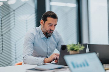 A professional man with a beard concentrates on his laptop in a contemporary office. A small plant...