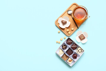 Delicious chocolate candies in box, wooden board, cup of tea, spoon and nuts on blue background