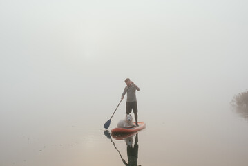 young man with tattooed arms on a sup board with his samoyed dog at the lake during sunset