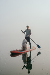 young man with tattooed arms on a sup board with his samoyed dog at the lake during sunset