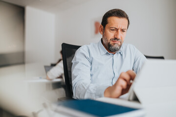 A professional man sits at a desk in a bright, modern office, concentrating on a tablet. The calm...
