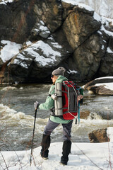 Caucasian young adult man standing on snowy riverbank holding trekking pole and wearing large...