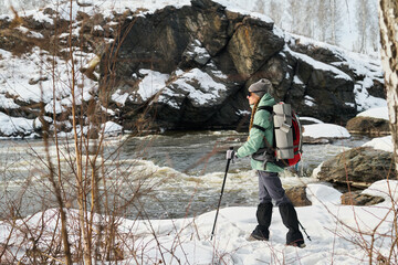 Caucasian young adult woman hiking along snowy riverbank carrying large backpack and trekking...