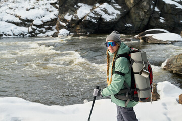 Caucasian young adult woman hiking along snowy riverbank carrying large backpack with camping gear,...