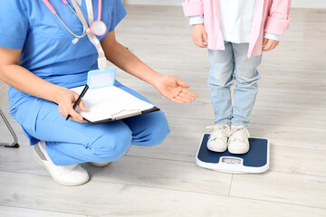 Female pediatrician with little girl measuring weight in clinic, closeup