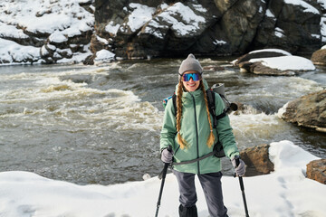 Caucasian young adult woman hiking with trekking poles near snowy riverbank, wearing outdoor gear...