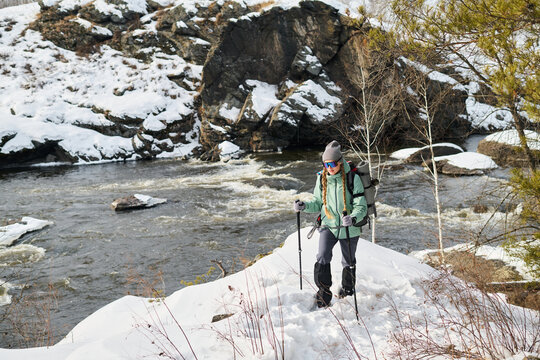 Caucasian young adult woman hiking along snowy riverbank carrying backpack and trekking poles, wearing winter gear and sunglasses, exploring winter landscape near flowing water - Powered by Adobe