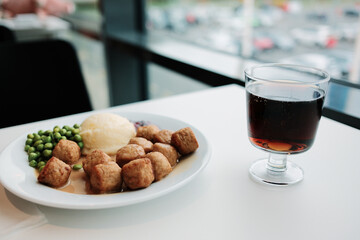 Plate of Swedish meatballs with mashed potatoes, green peas, and gravy served beside a glass of soda on a white table by the window