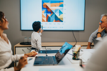 A young presenter stands before a large screen displaying a vibrant geometric chart while colleagues observe, laptops open, creating a collaborative and modern office atmosphere.