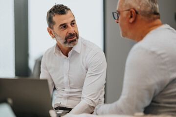 Fototapeta premium Two men in a modern office talk beside a laptop, sharing ideas and planning a project. The scene conveys collaboration, focus, and professional teamwork.