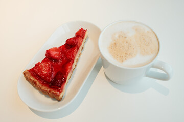 Slice of glazed strawberry tart with golden crust served beside a cup of cappuccino on a clean white surface. The concept of breakfast in a cafe