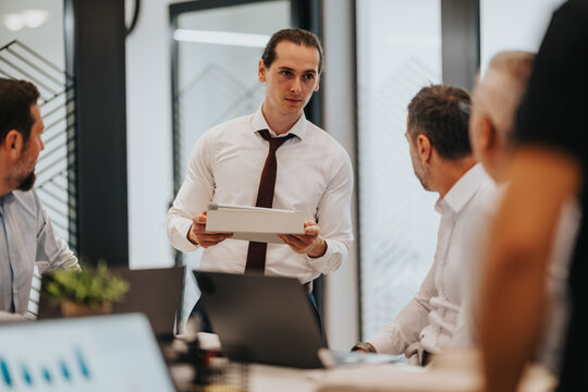 A team gathers in a contemporary office for a meeting. One man stands with a tablet, presenting to colleagues around a conference table with laptops, creating a collaborative, focused atmosphere.