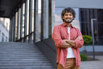 Portrait of a smiling young Indian man in a red shirt standing confidently with his arms crossed on his chest, looking at the camera